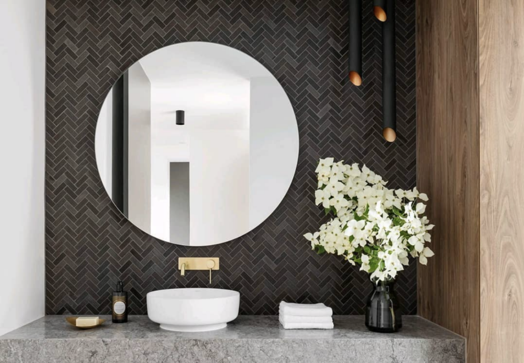 Modern bathroom vanity with stone bench top, dark tiling, round mirror and vase of white flowers