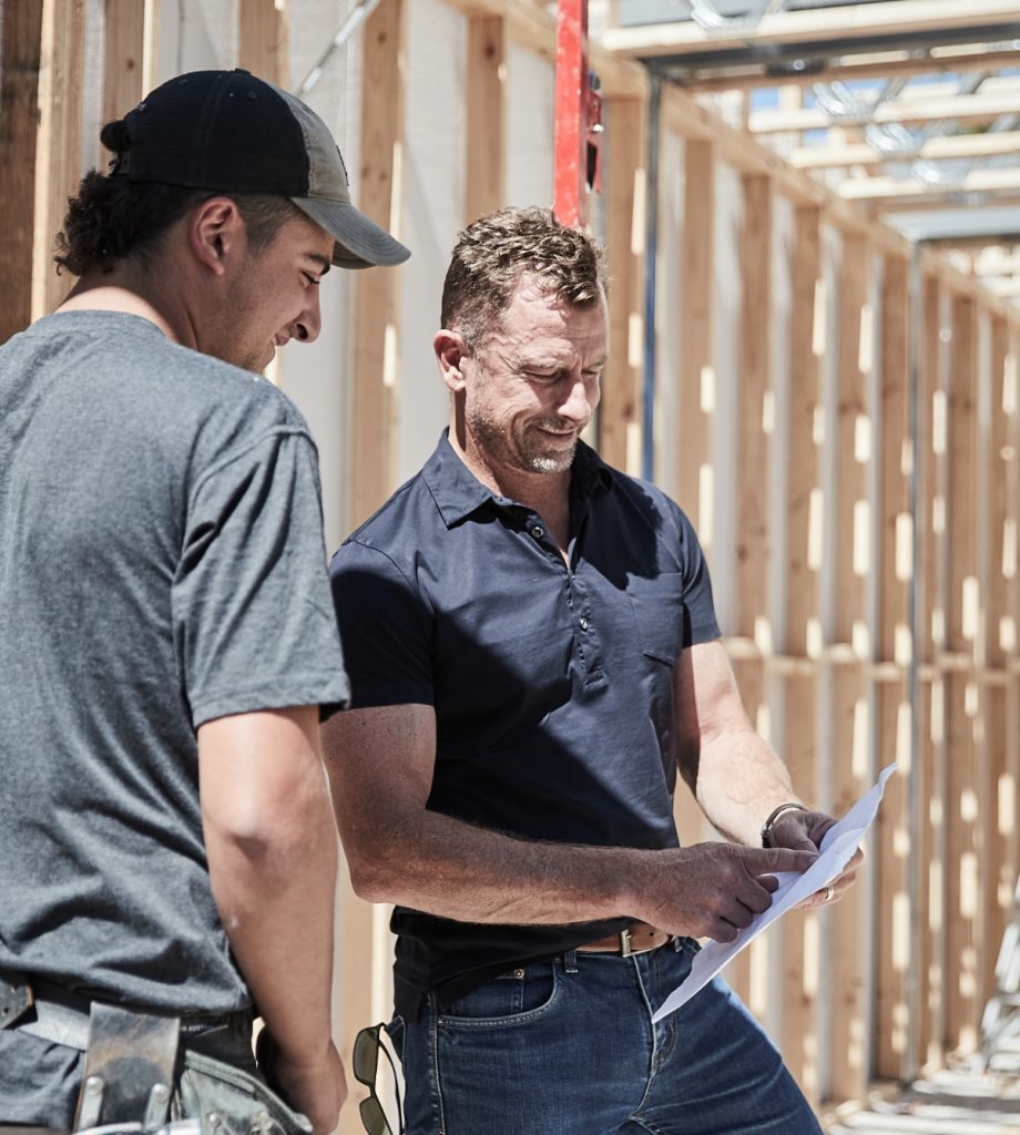 Two men standing next to timber framing on a building site looking at plans