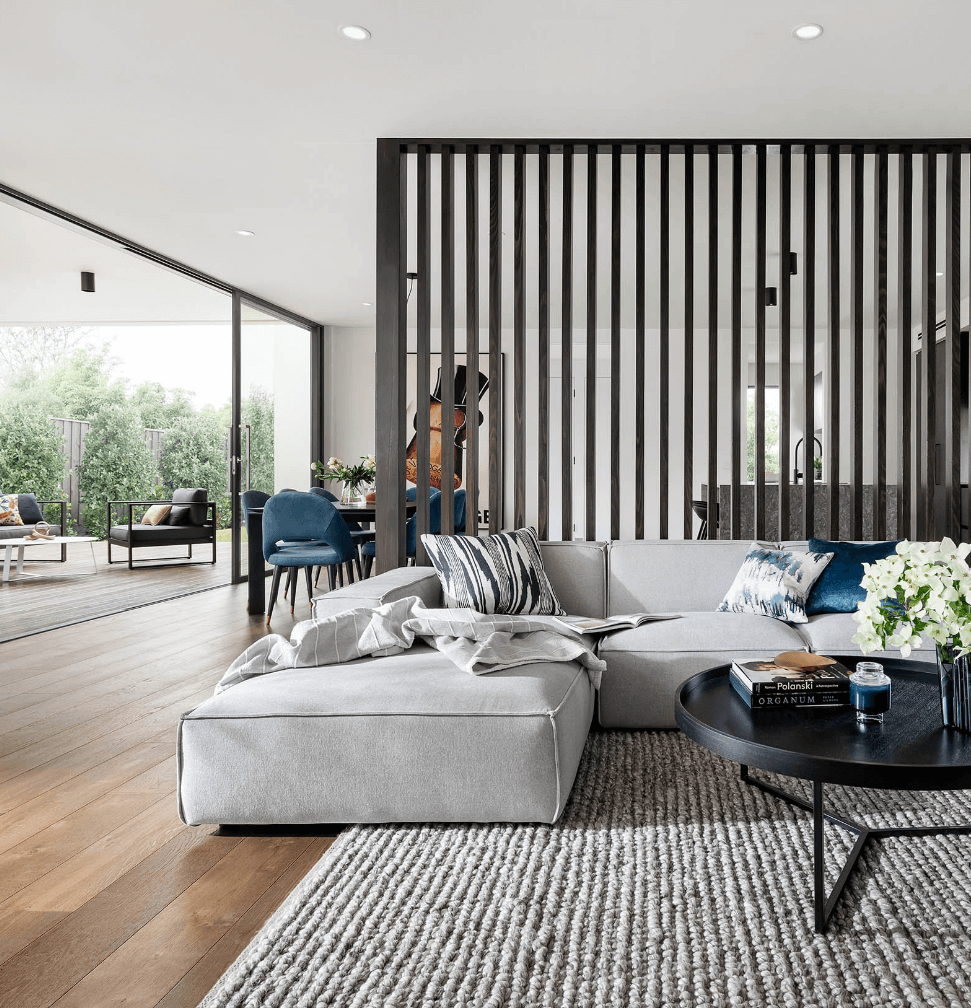 Modern living area with large grey couch and black coffee table, and room divider through to dining and kitchen