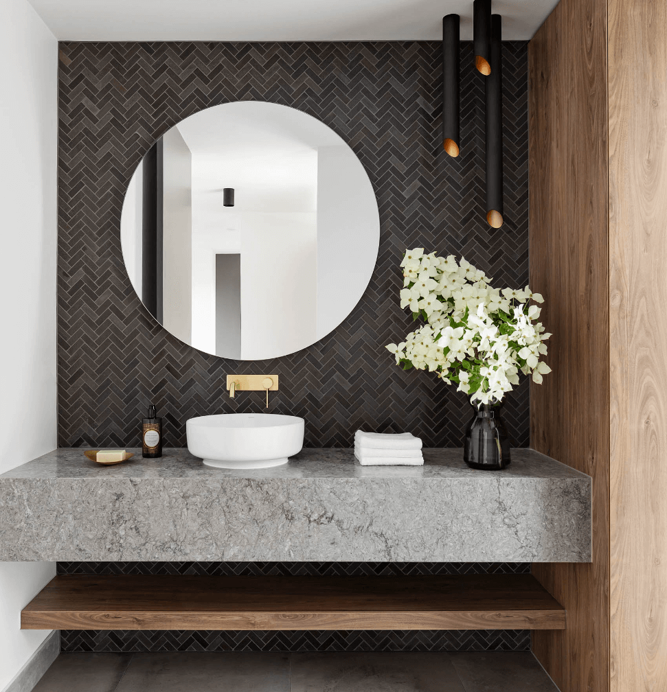 Modern bathroom vanity with stone bench top, dark tiling, round mirror, and vase of white flowers