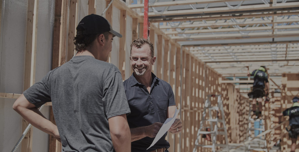 Two men on standing next to timber framing on building site, looking at plans