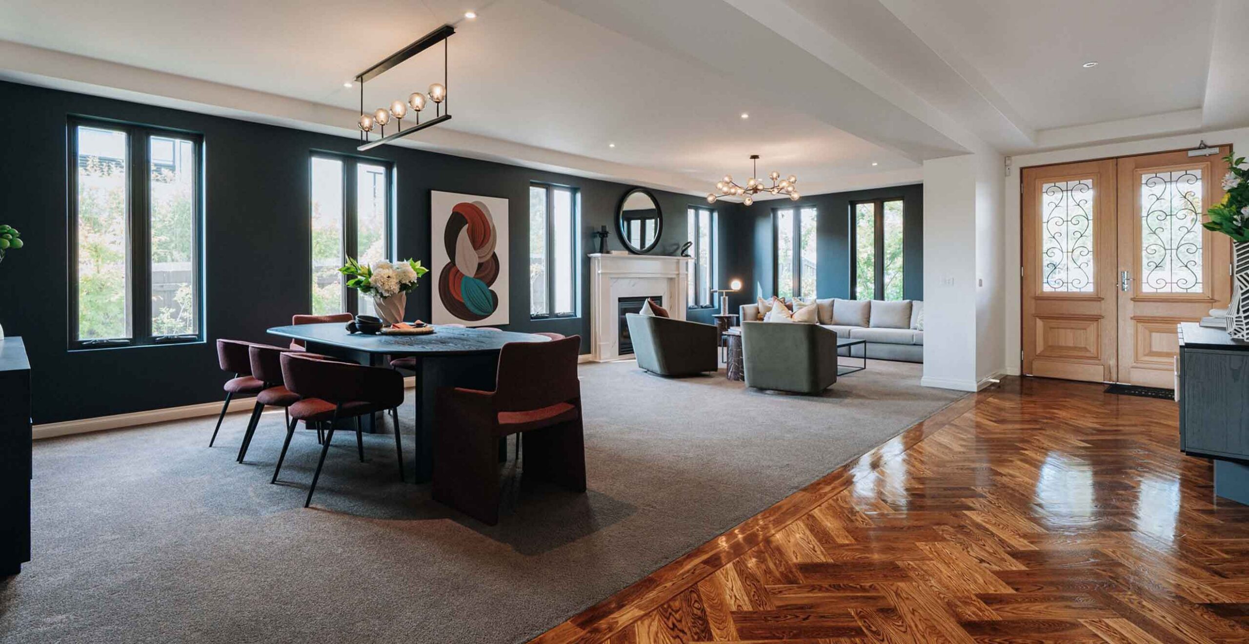 Open-concept living and dining room with dark accent wall, large windows, and a round painting; dining table with red chairs and a modern chandelier above.