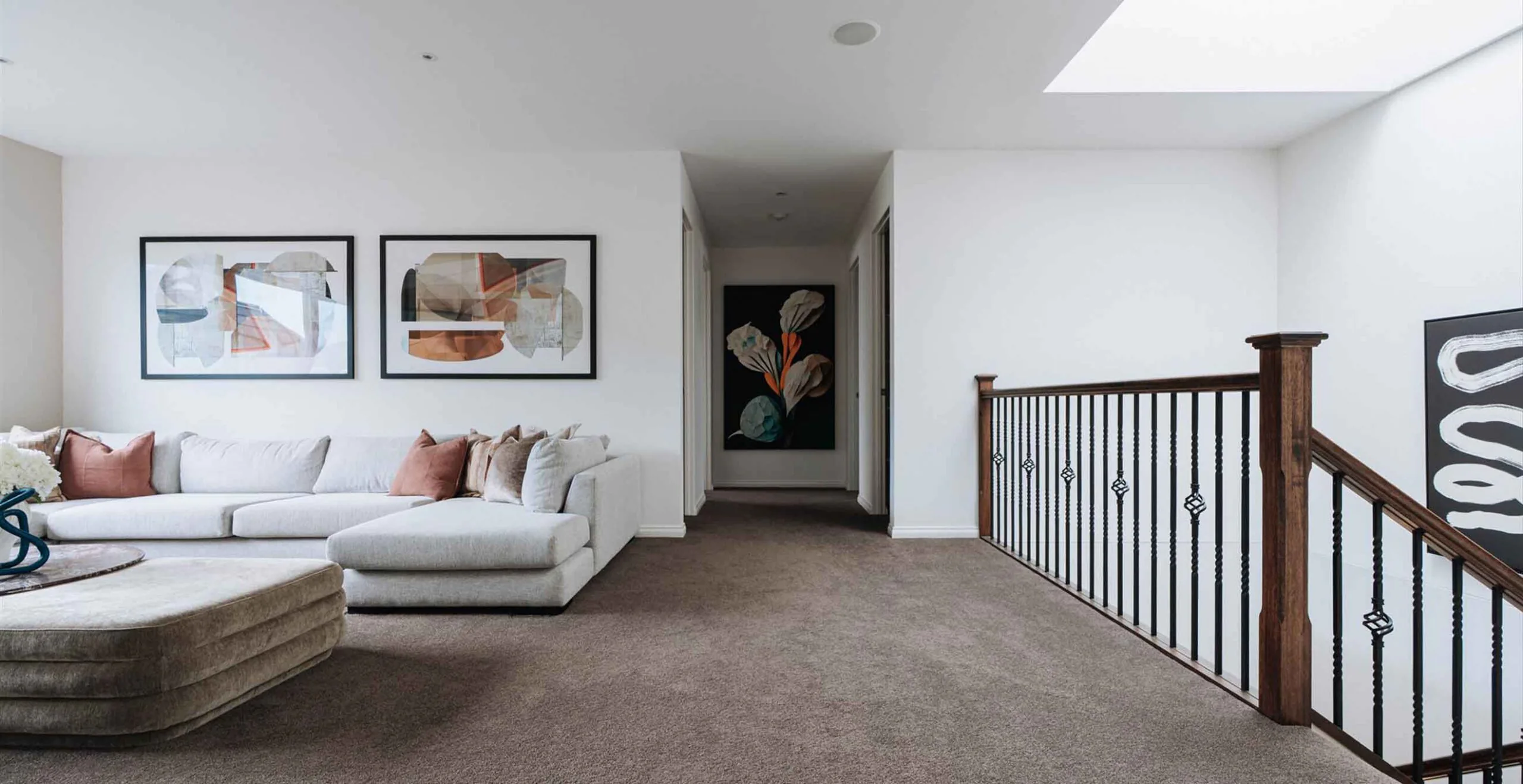 Bright living room with a light gray sectional sofa, pink/beige cushions, and two abstract framed artworks on the wall; a hallway leads to a dark artwork at the end, with a wooden railing on the right.