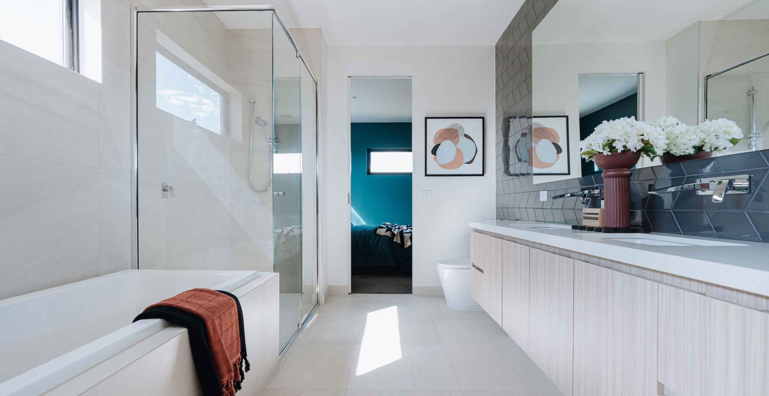 Bright modern bathroom with a white bathtub on the left, glass shower enclosure, and a long wooden vanity with a white countertop and white flowers in a red vase close to the mirror.