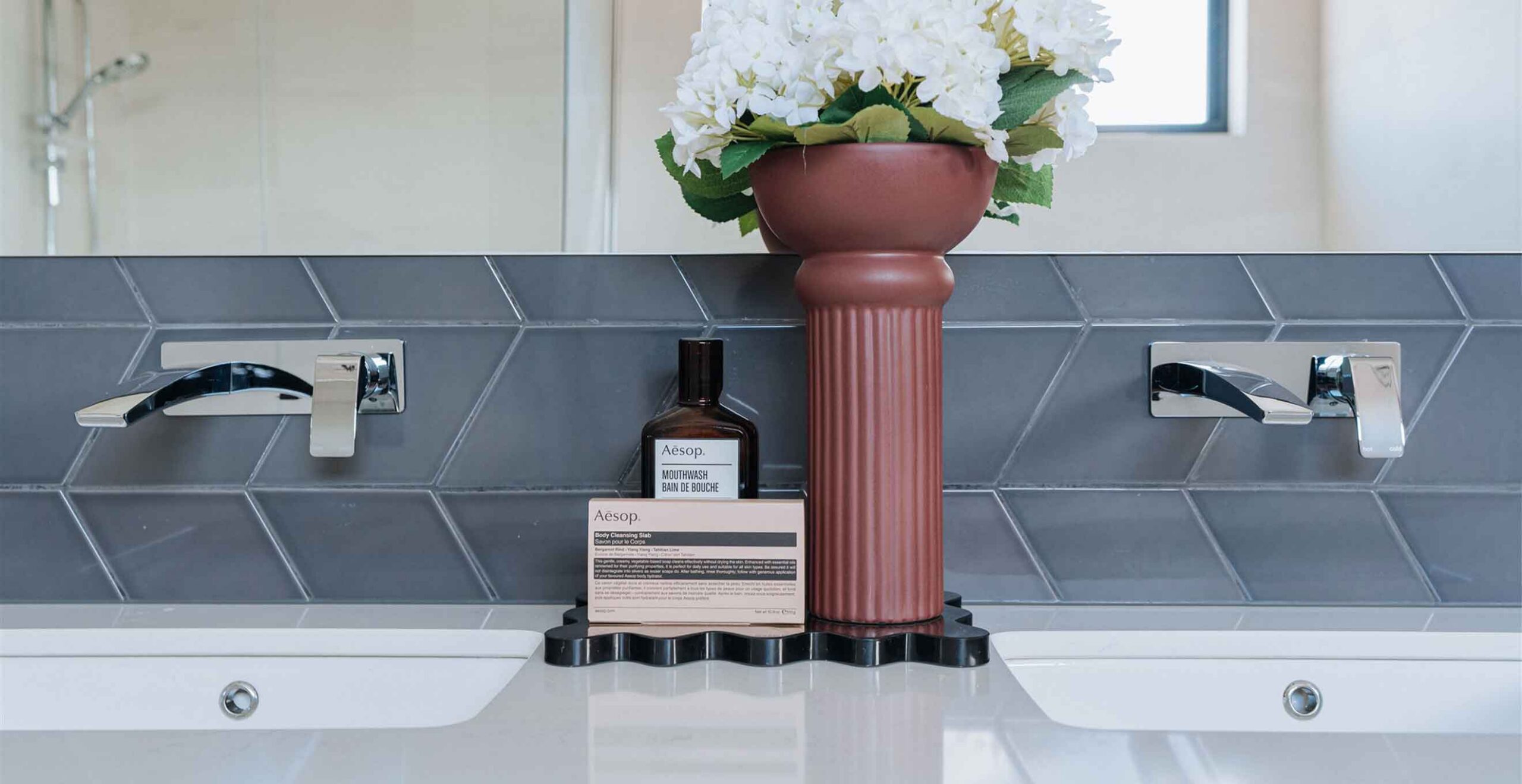 Symmetric bathroom vanity with two chrome faucets, a pink ribbed vase of white flowers, and an Aesop hand wash bottle nearby.