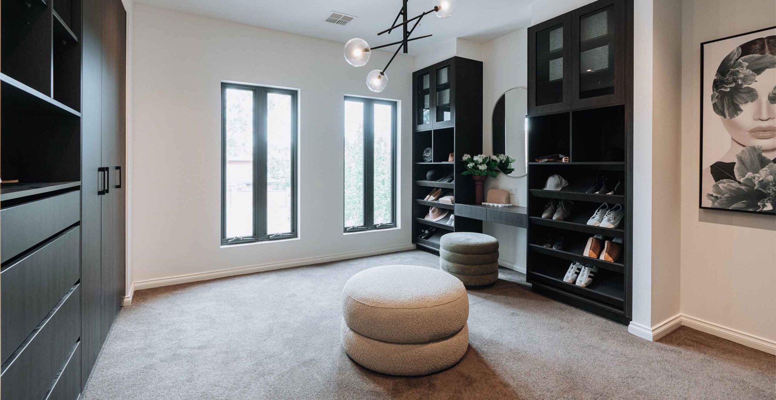 Luxurious walk-in closet with black built-in cabinets, open shoe shelves, and two round beige ottomans on a carpeted floor.