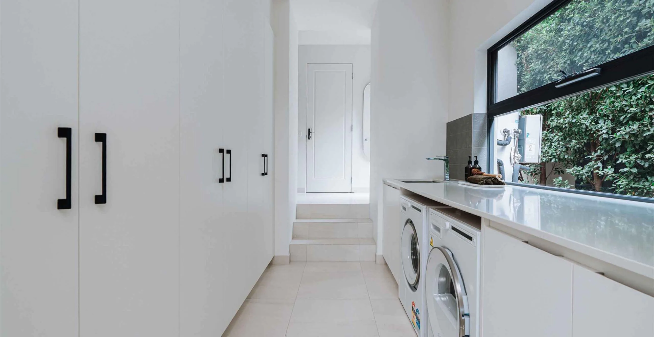 Bright laundry room with tall white cabinets on the left, a glossy countertop, and front-loading washer and dryer under the windowed wall on the right.