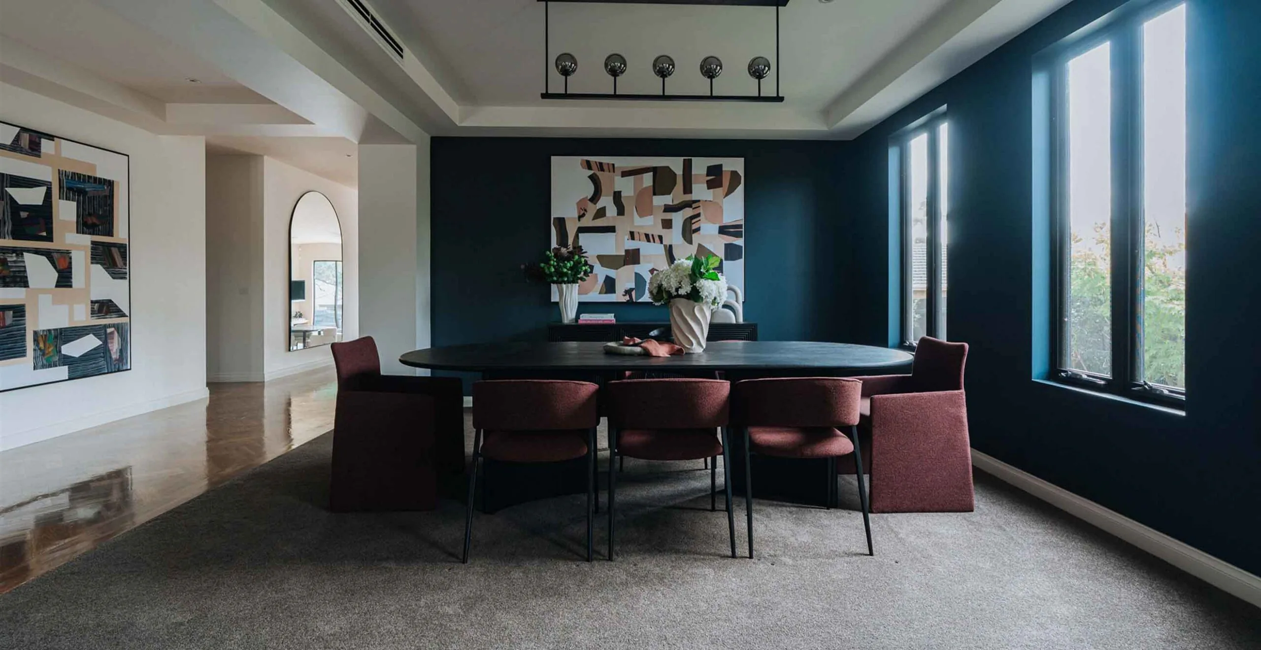 Modern dining room with a round black table, burgundy chairs, and a dark blue accent wall featuring a large abstract canvas center stage.