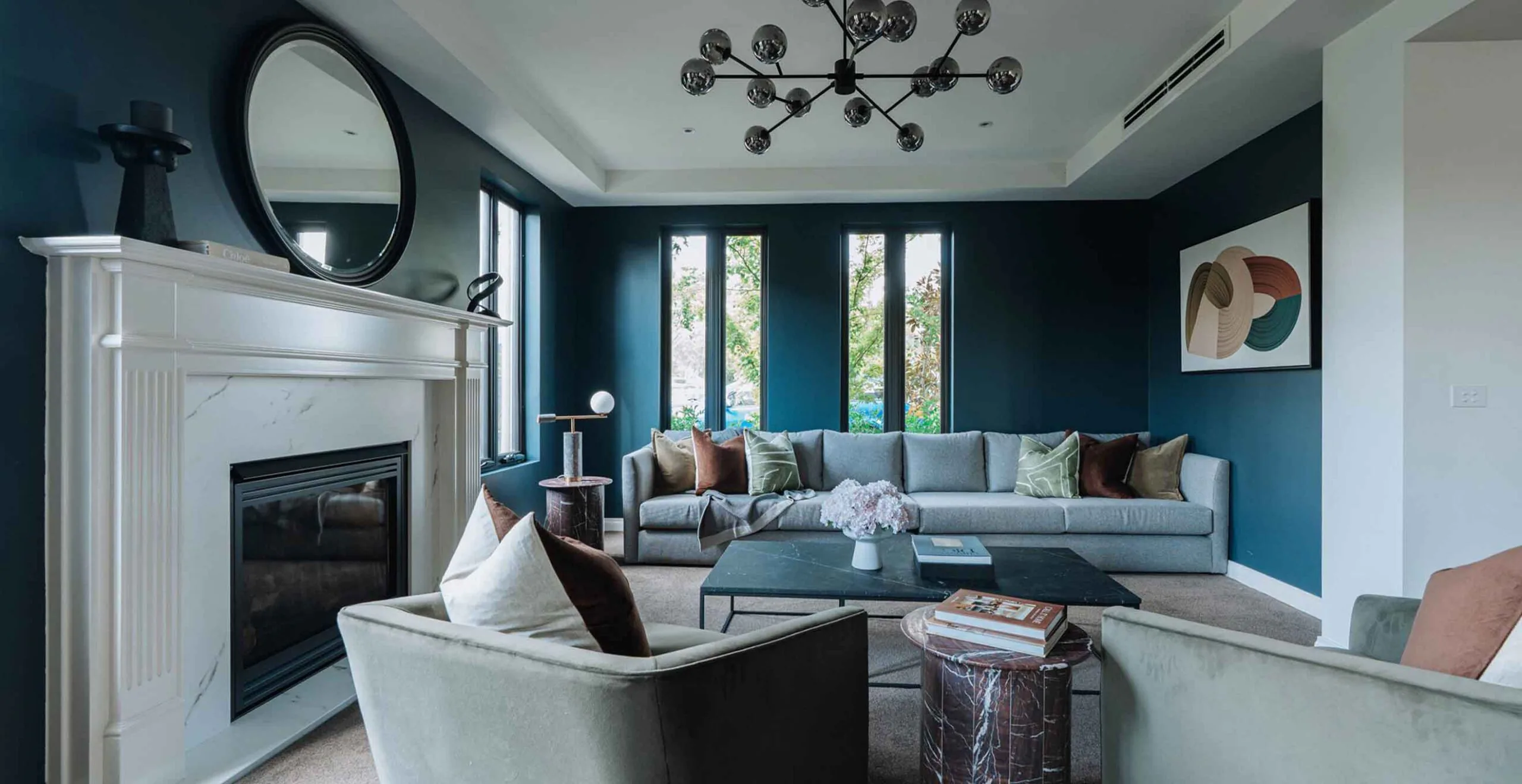Modern living room with a white marble fireplace, circular mirror, and teal walls above a gray sectional sofa.