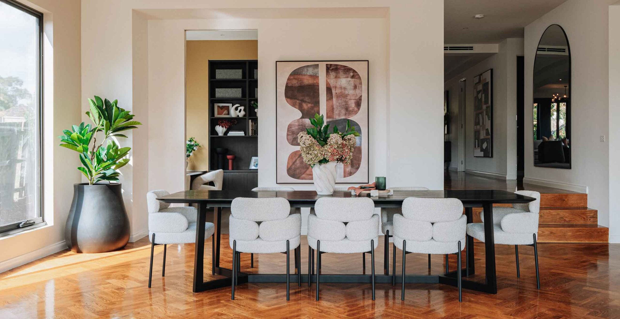 Contemporary dining room with a dark rectangular table and light gray upholstered chairs, centered by a white vase of flowers and a large abstract wall art piece.