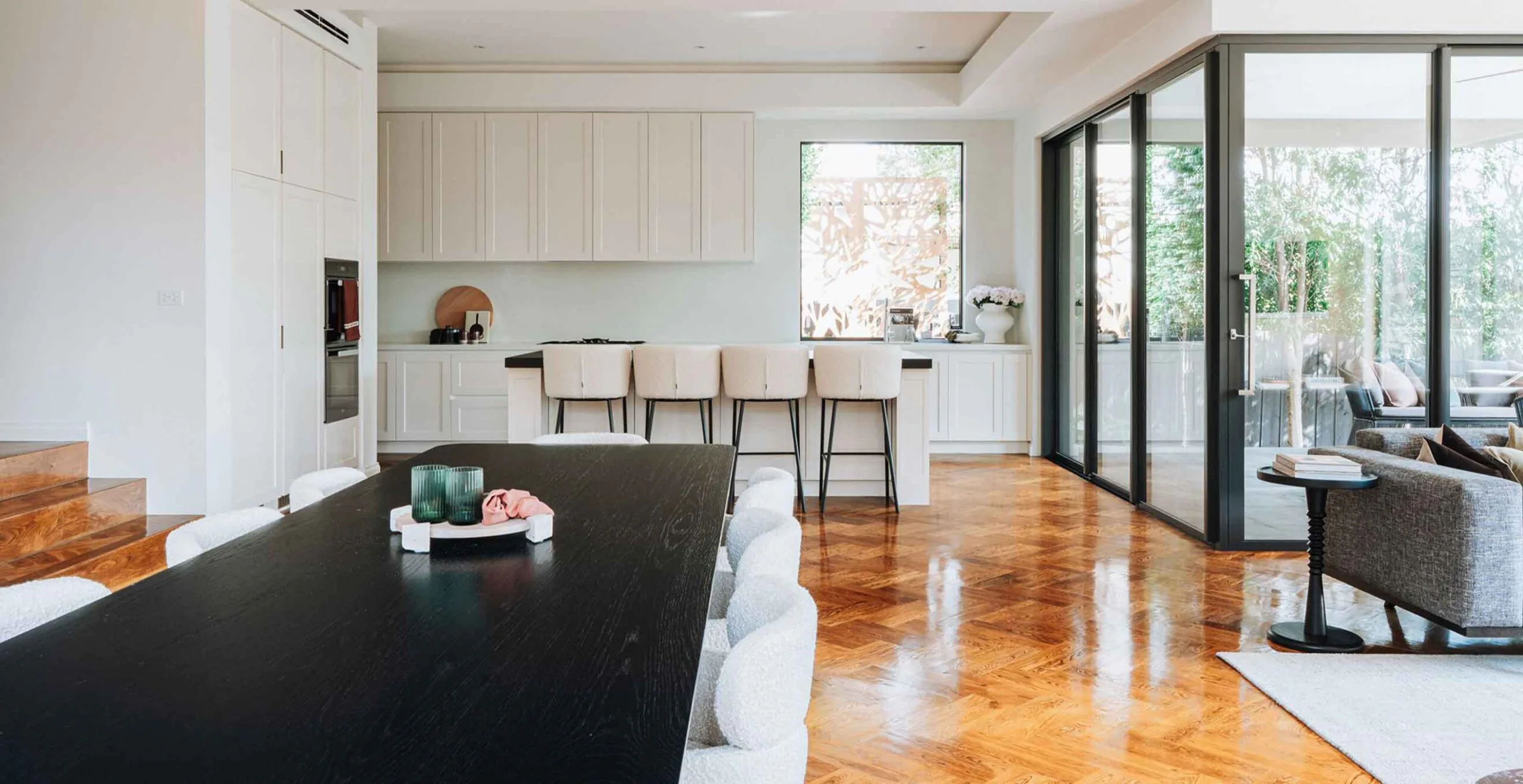 Open-concept kitchen with white cabinets and an island with beige bar stools facing a large windowed wall and outdoor patio doors.