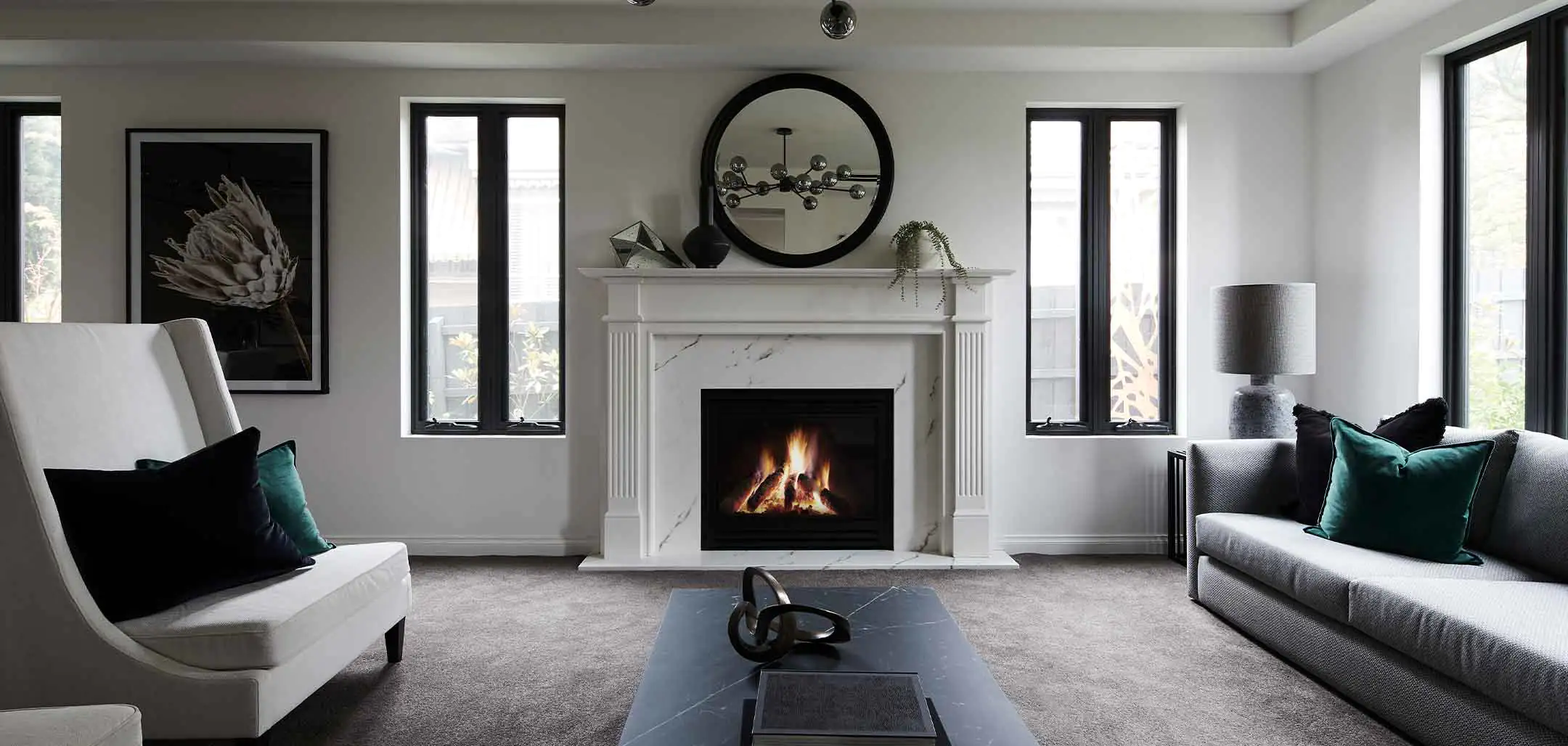 Modern living room with a lit fireplace, marble mantel, and round black-framed mirror above; two gray sofas facing a dark coffee table.
