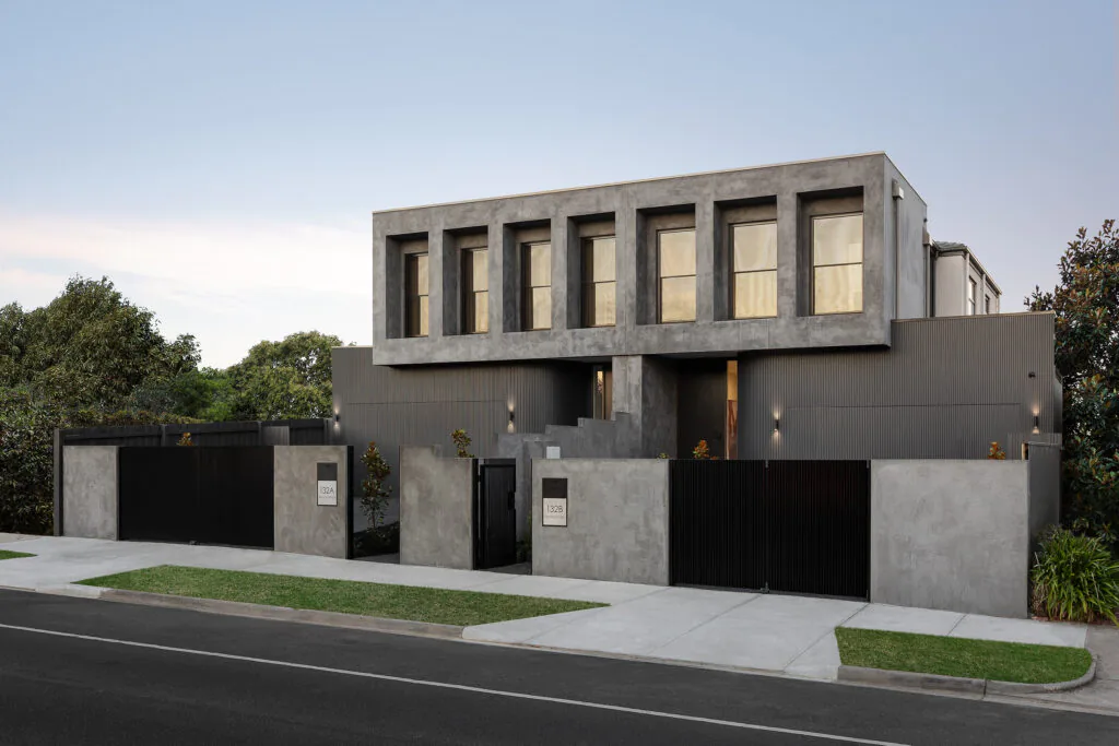Modern two-story concrete house with large rectangular windows and a tall black metal fence beside a sidewalk.