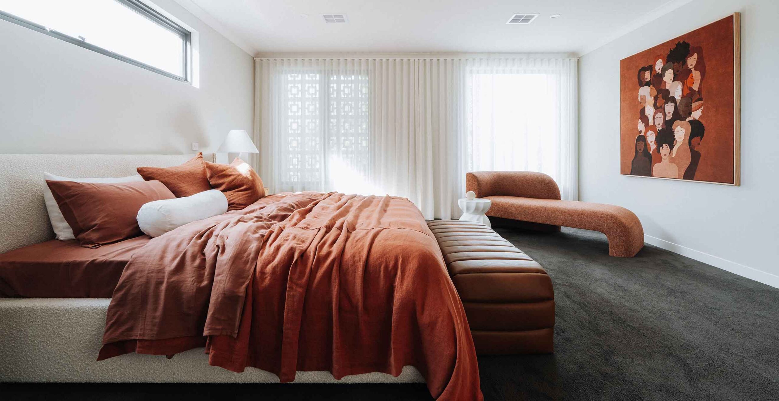 Bedroom with orange duvet, white headboard, curved orange chaise, and a large multi-portrait artwork on the right wall.