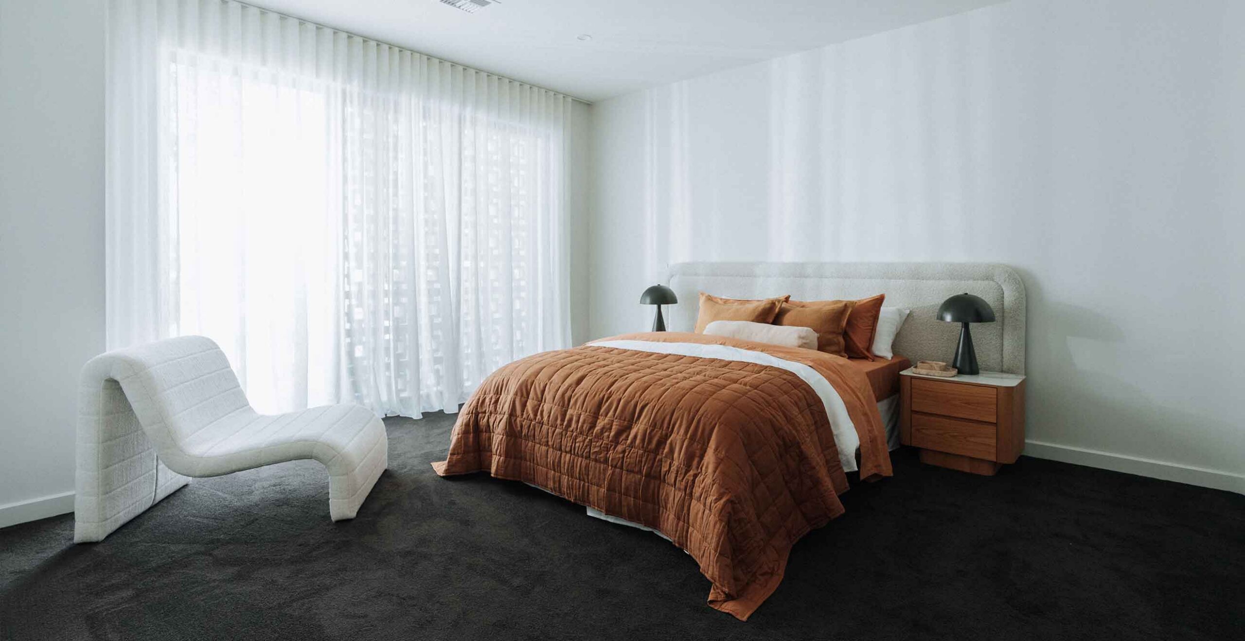 Bedroom with an orange quilt on a white bed, black bedside lamps, wooden nightstands, and a white curved chair by large sheer-curtained windows.
