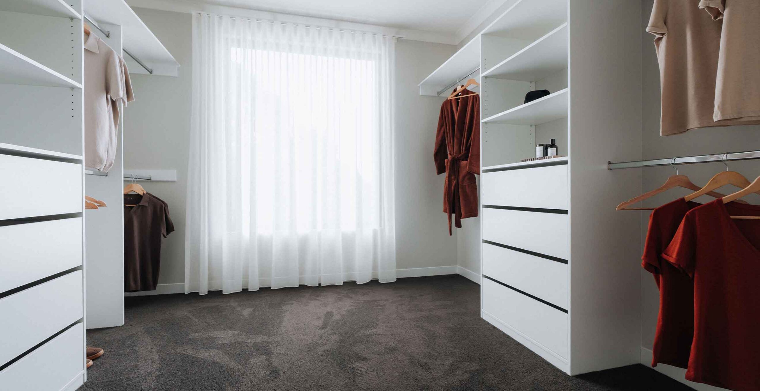 Walk-in closet with white built-in shelves and hanging rods, beige and burgundy robes and clothing hanging neatly.