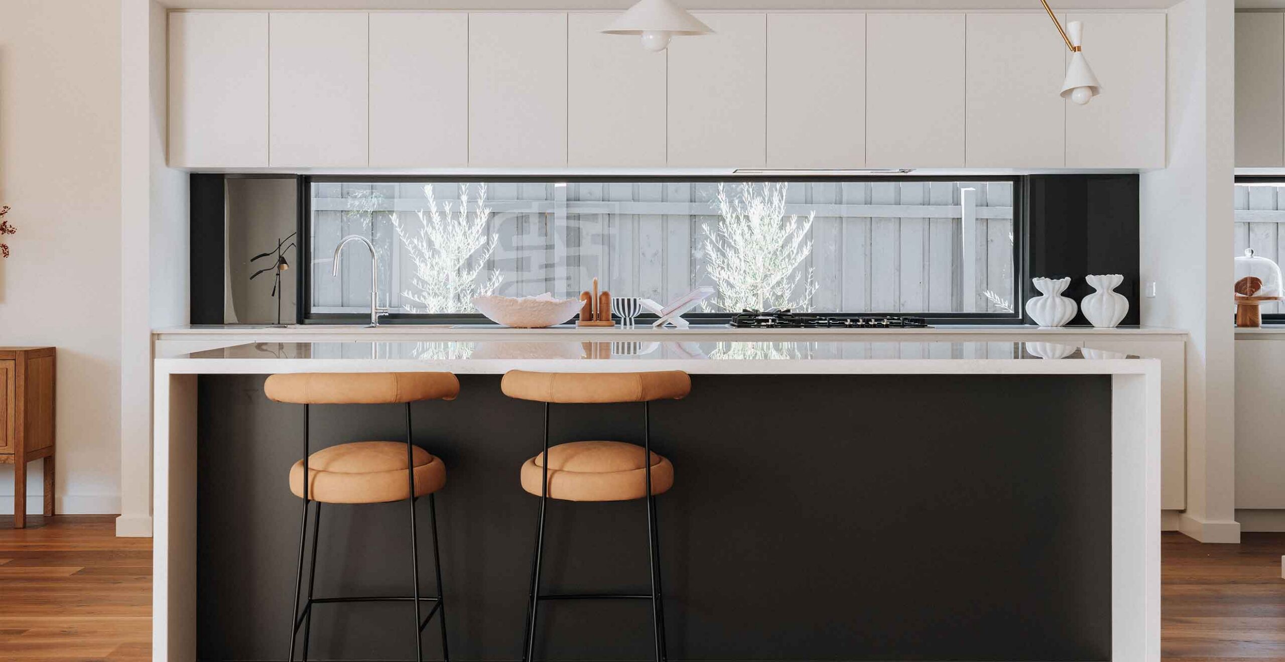 Modern white kitchen with a marble-topped island, tan bar stools, and a large window behind the sink area.