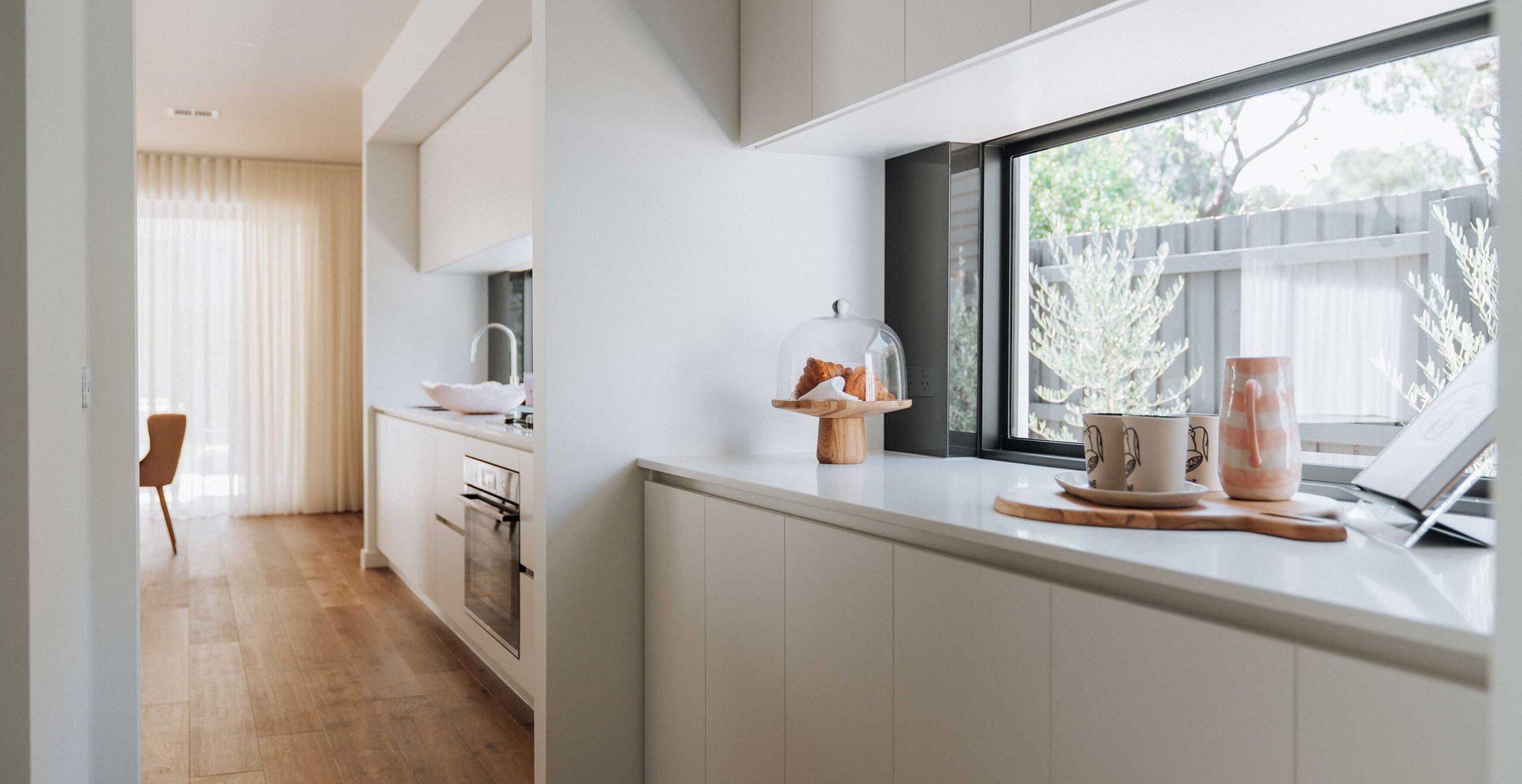 Modern kitchen with white cabinets, hardwood floor, and a pastry under a glass cloche on the counter by a large window.