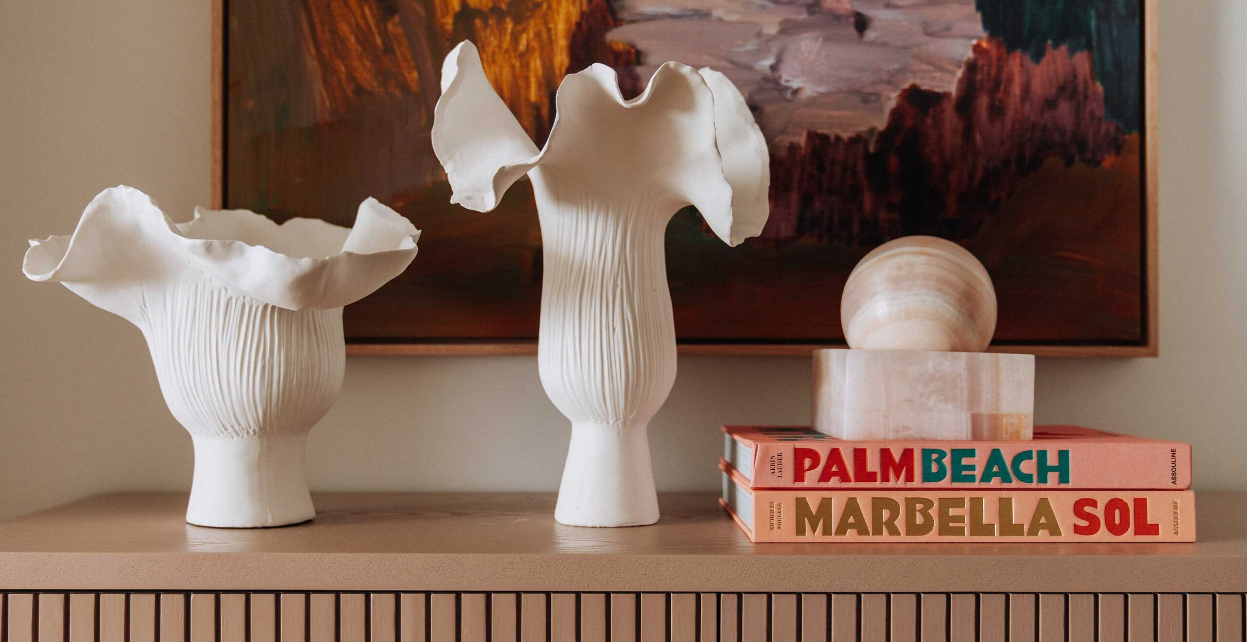 White ceramic vases with ruffled, wide rims on a shelf beside a round stone sculpture and colorful book spines reading Palm Beach and Marbella Sol under a painting.