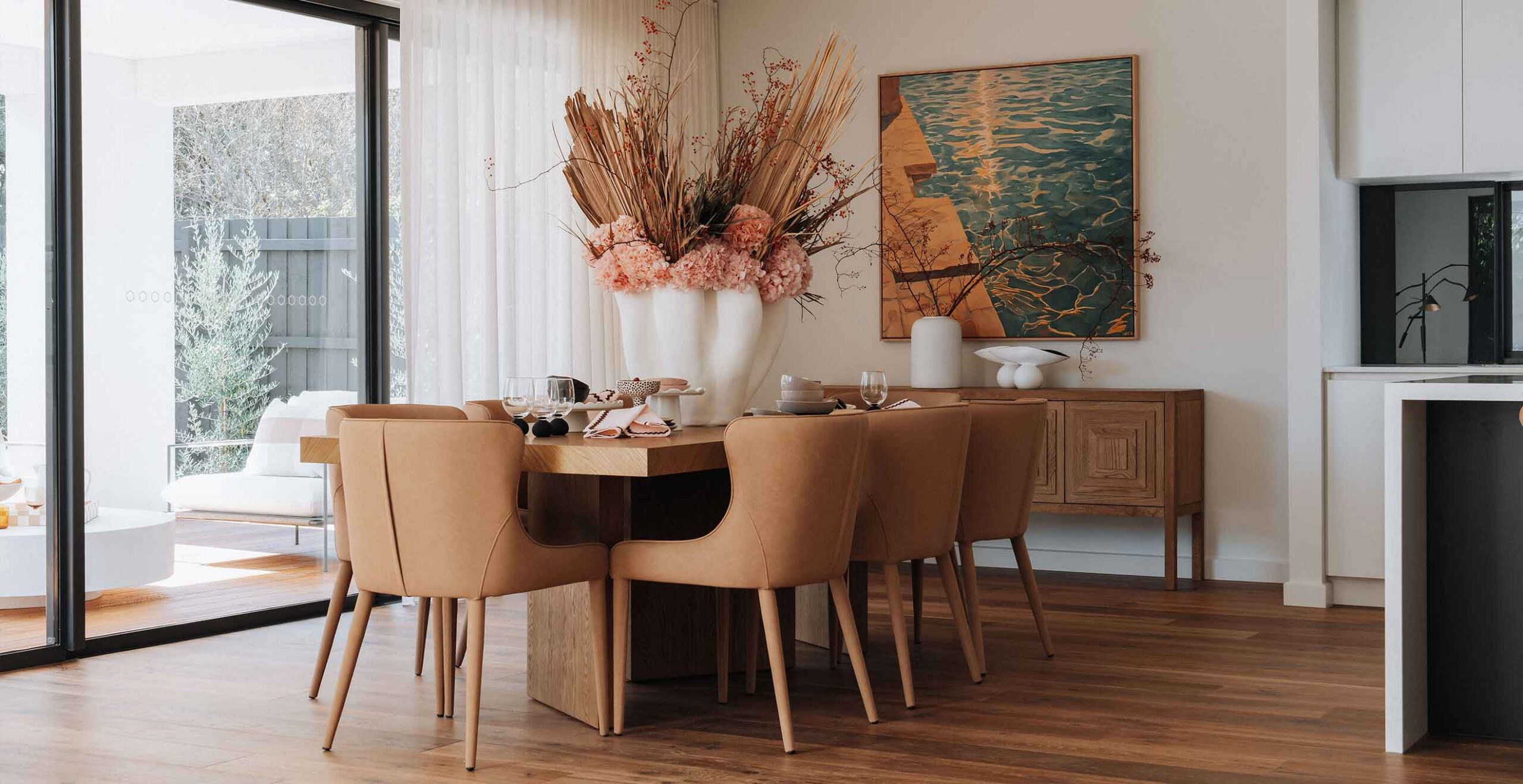 Modern dining room with a wooden table, tan chairs, and a tall white vase of pink flowers as a centerpiece.