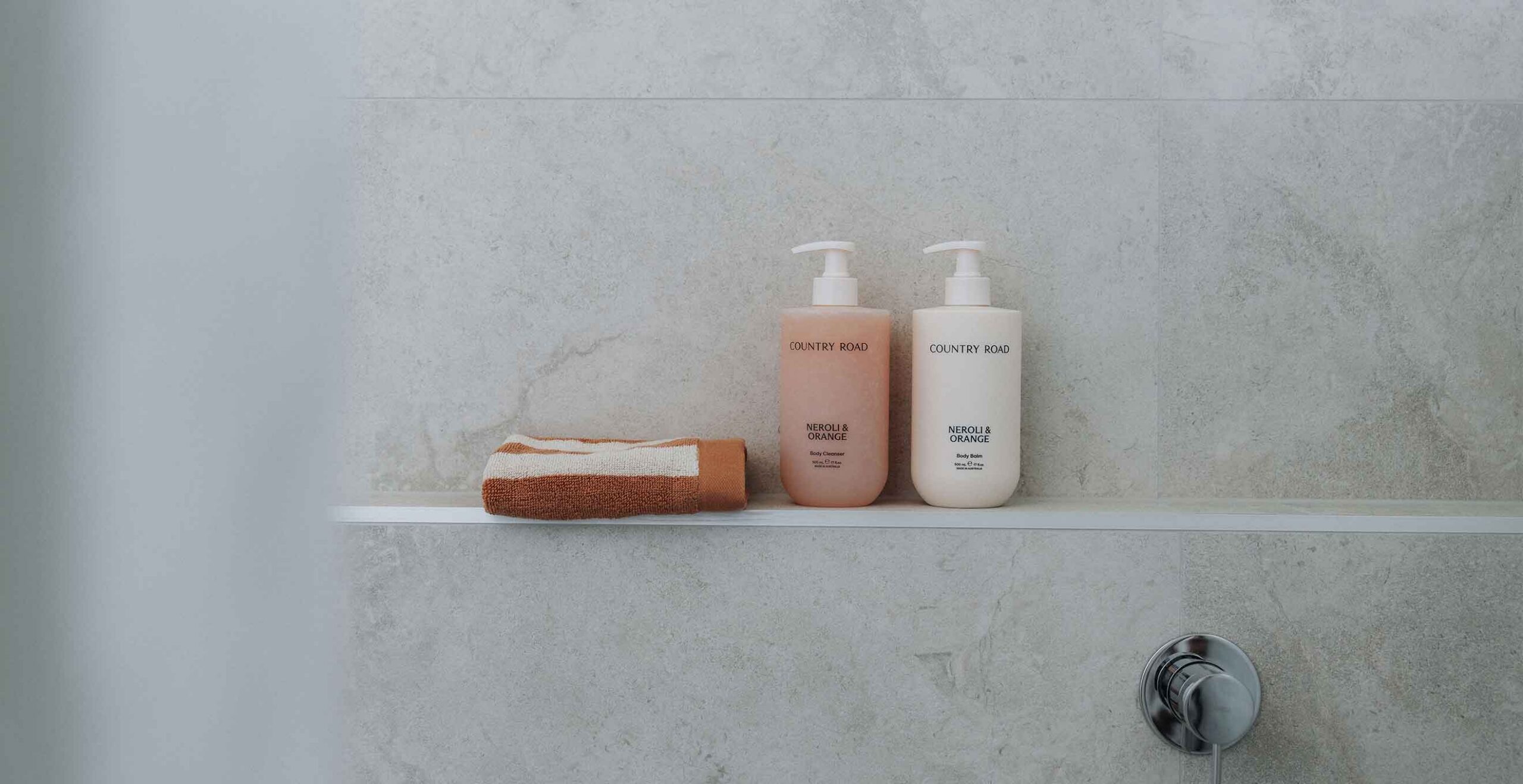 Two Country Road pump bottles (neroli & orange) on a bathroom shelf next to a folded orange and white towel, with a gray tiled wall background.