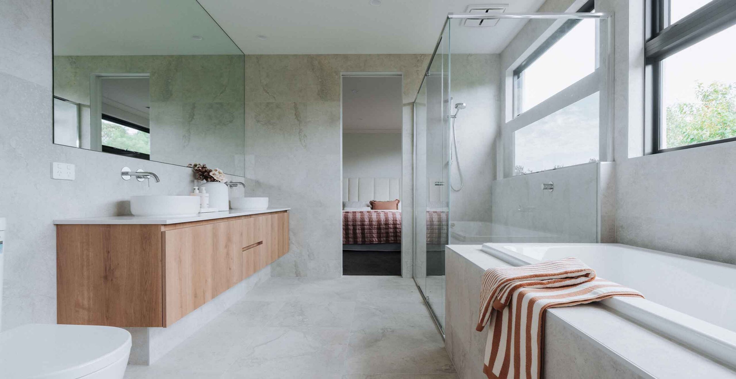 Modern bathroom with a wooden double-sink vanity, marble walls, and a glass shower; view into a bedroom through an open doorway in the back.