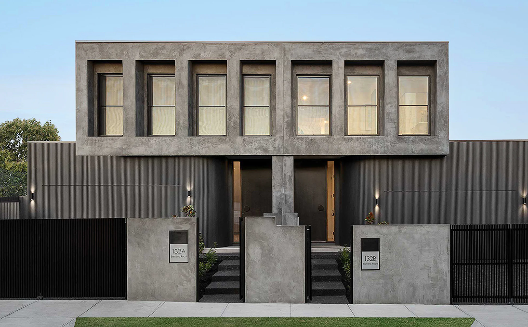 Modern gray concrete building facade with six tall rectangular windows and two entry doors beneath a cantilevered upper level. (Addresses 132A and 132B)