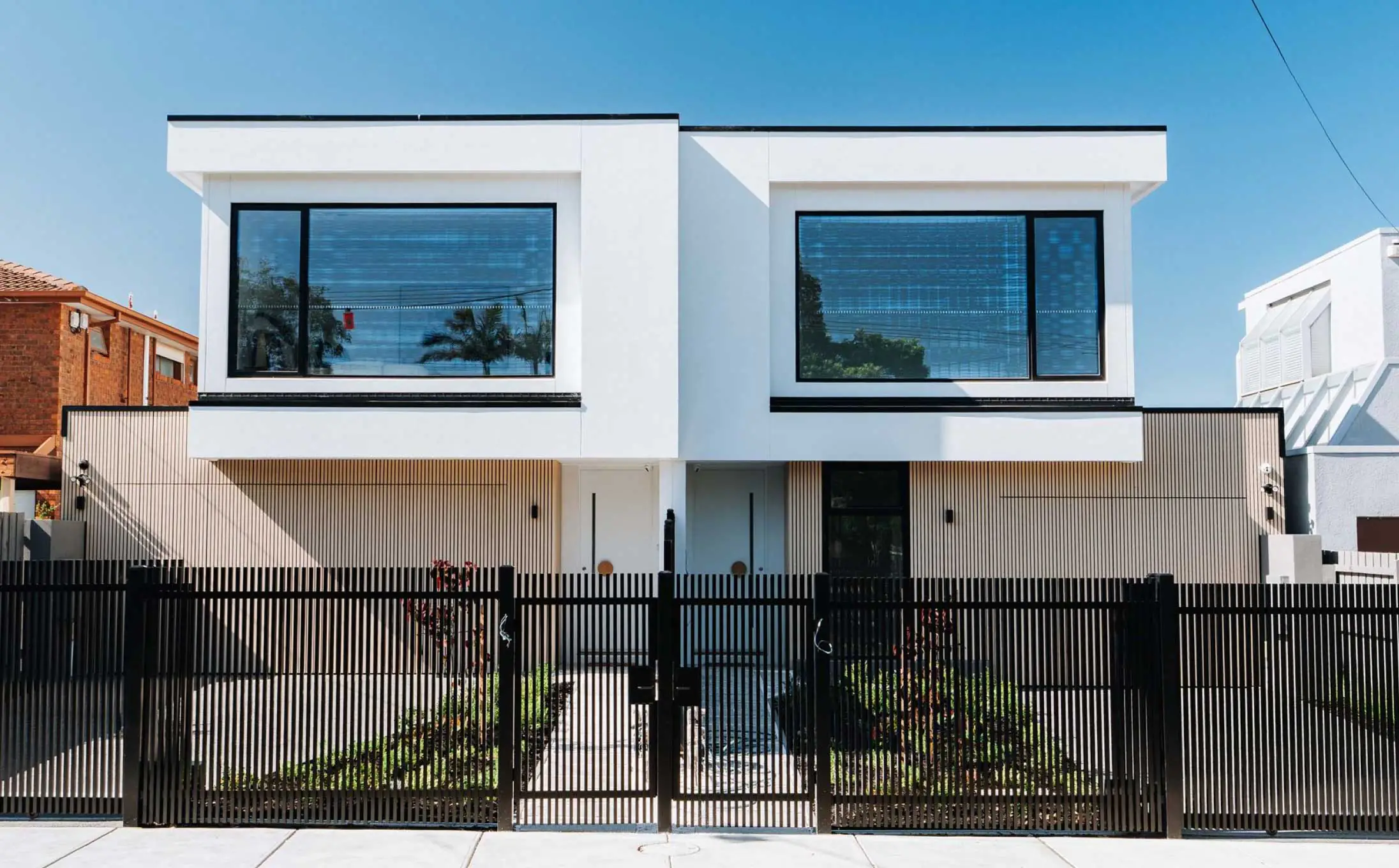 Modern white two‑story house with large black-framed windows, metal fencing, and a gated front yard under a clear blue sky.
