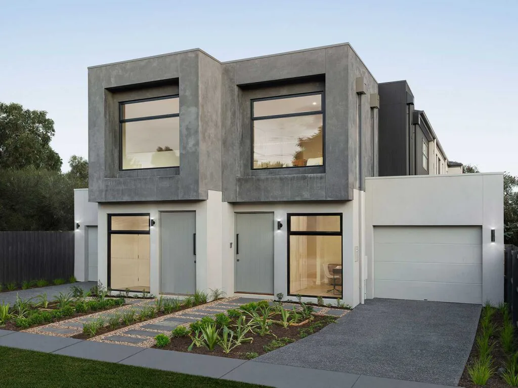 Modern two-story house with concrete gray façade, large front windows, and a double garage; landscaped front yard with gravel paths and greenery.