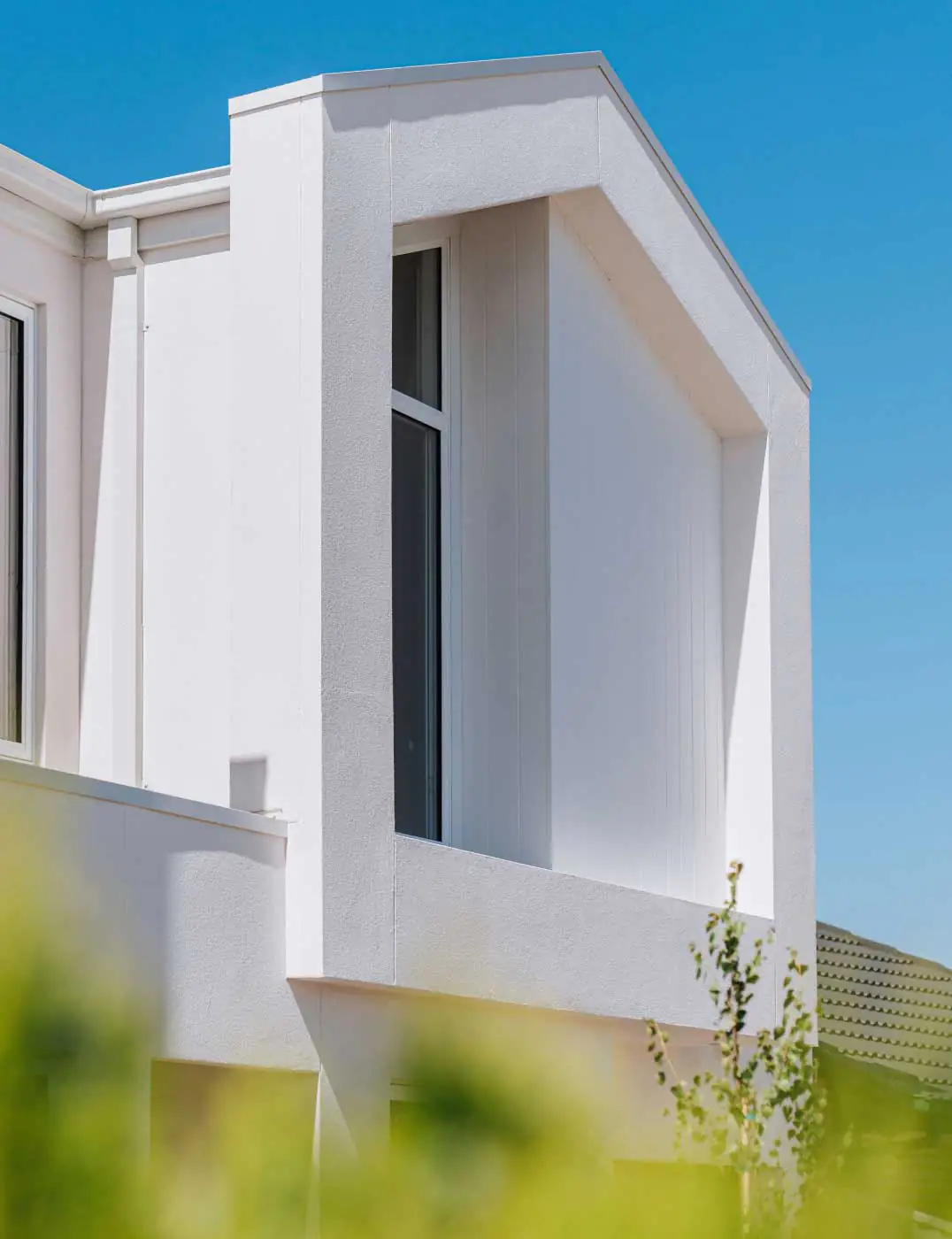 White modern building corner with angular window alcove against a bright blue sky, blurred greenery in foreground