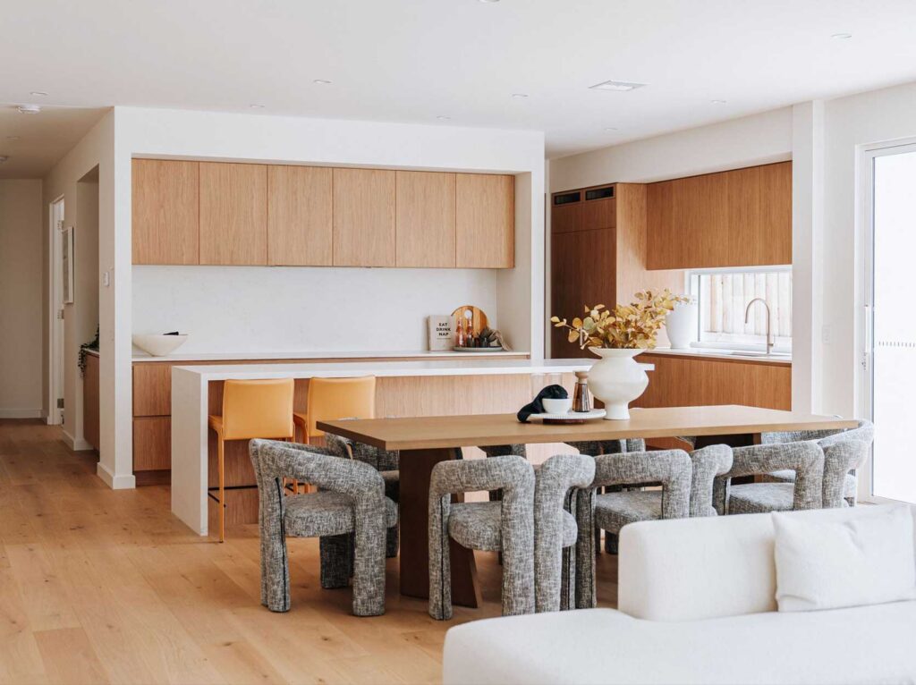 Open-plan kitchen and dining area with light oak cabinets, white countertops, and a long wooden table with grey chairs.