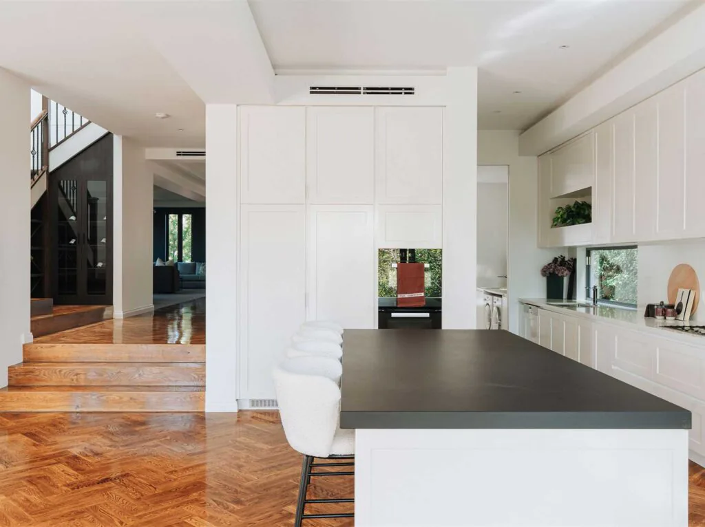 Open-concept kitchen with white paneled cabinets, a large dark island, and wooden herringbone floors; stairs and living area visible to the left.
