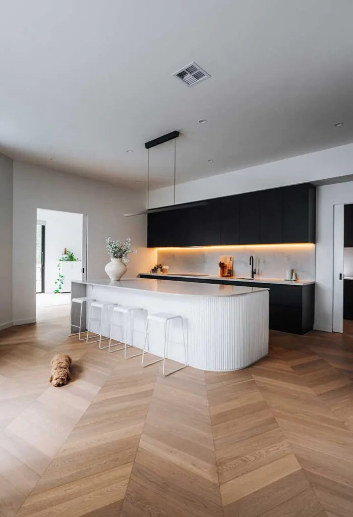 Open, minimalist kitchen with a curved white island and five white stools, black upper cabinets, warm under-cabinet lighting, and a small dog resting on light wood chevron flooring.