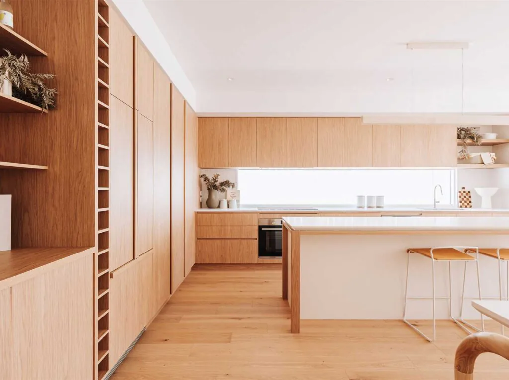 Bright minimalist kitchen with warm wood cabinets, a white island, and light wood flooring; open shelves and bar seating add a cozy, modern touch.