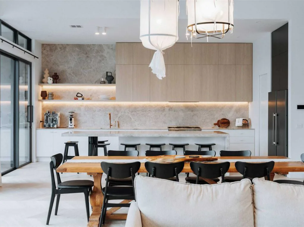 Open-concept kitchen and dining area with a long wooden table, black chairs, and beige cabinetry under warm lighting.