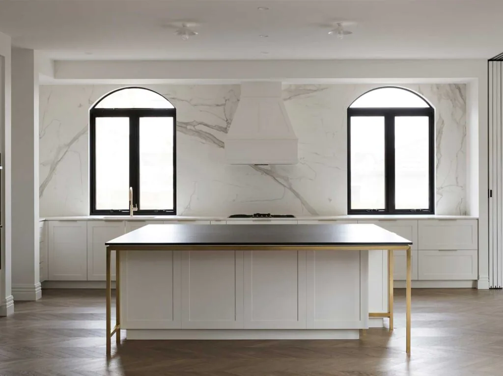 Modern white kitchen with marble backsplash, black-framed arched windows, and a central white island with gold legs.