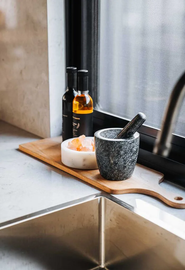 Stone mortar and pestle with salt rocks on a wooden board by a kitchen window, oil bottles in the background.