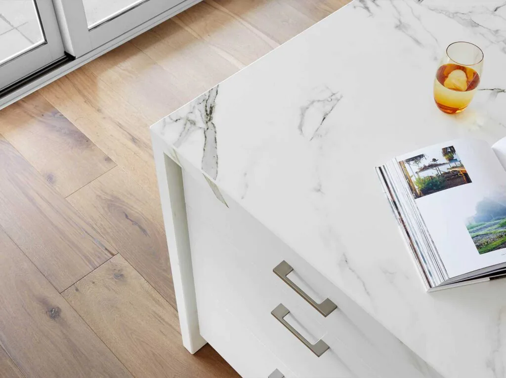White marble-topped cabinet with silver handles on a light wood floor, an open magazine resting on the surface, and a glass of amber drink nearby.
