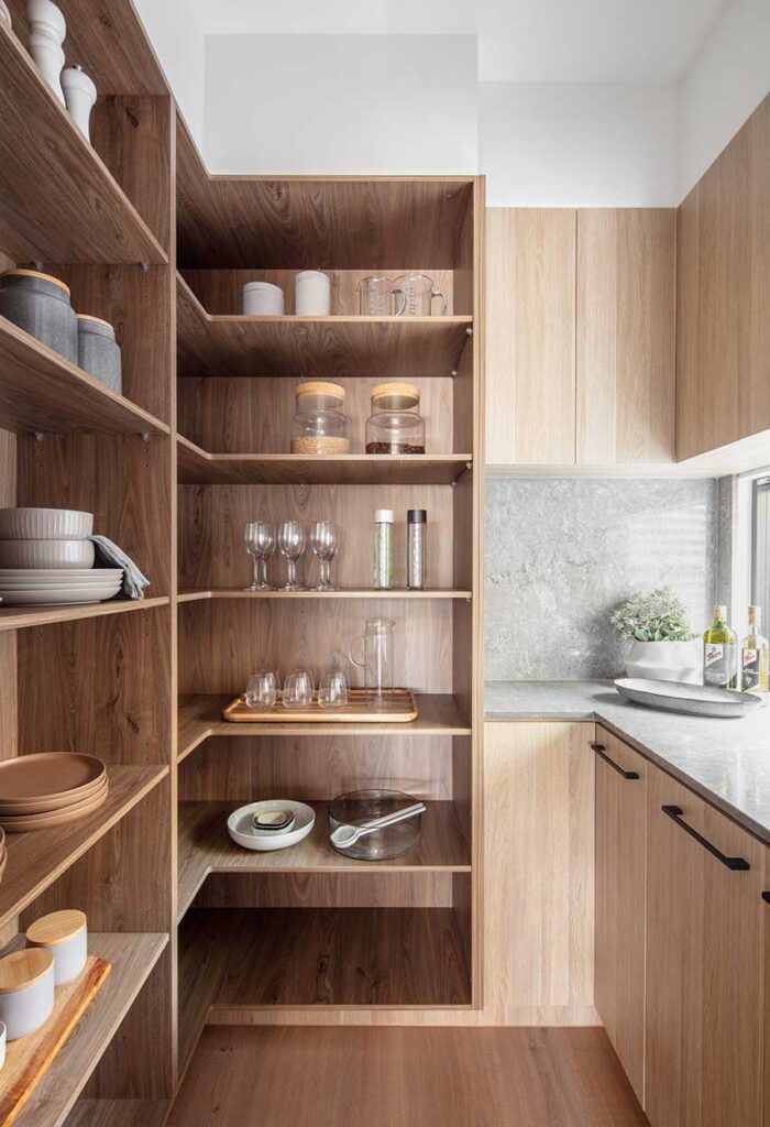 Open wooden pantry shelves displaying jars, glassware, plates, and bowls beside a light countertop in a modern kitchen.