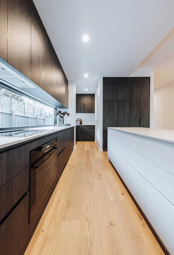 Modern galley kitchen with dark wood cabinetry on the left, a white island on the right, and warm hardwood flooring under recessed ceiling lights.
