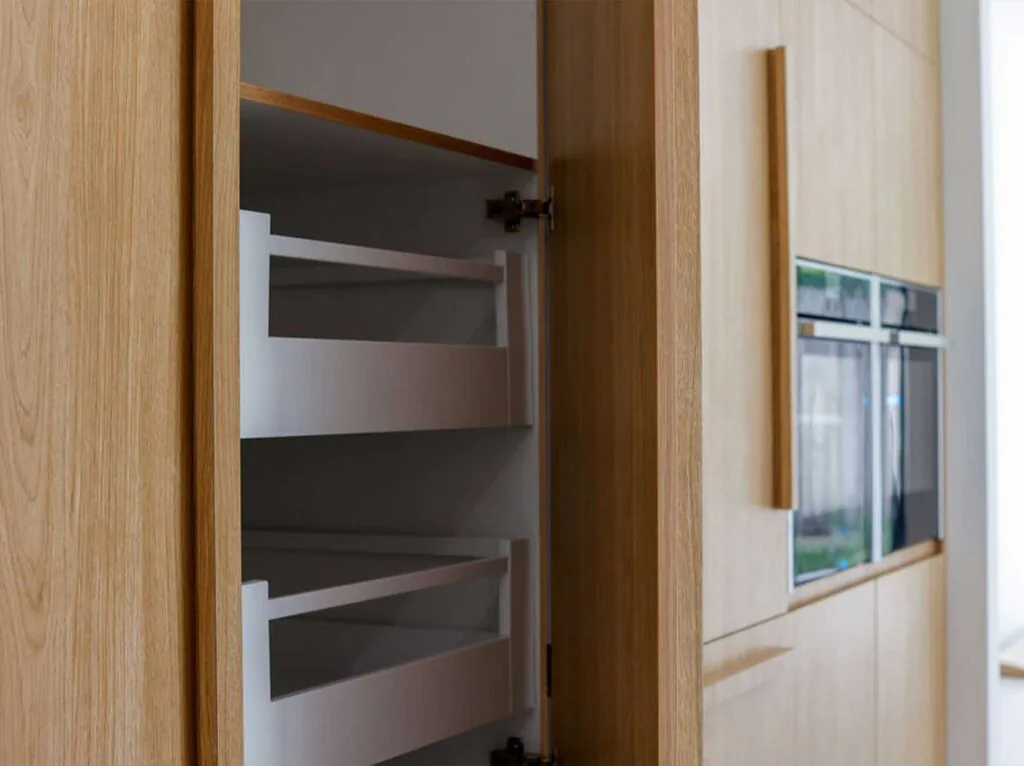 Open cabinet with white pull-out shelves next to a built-in oven in a wood kitchen setting.