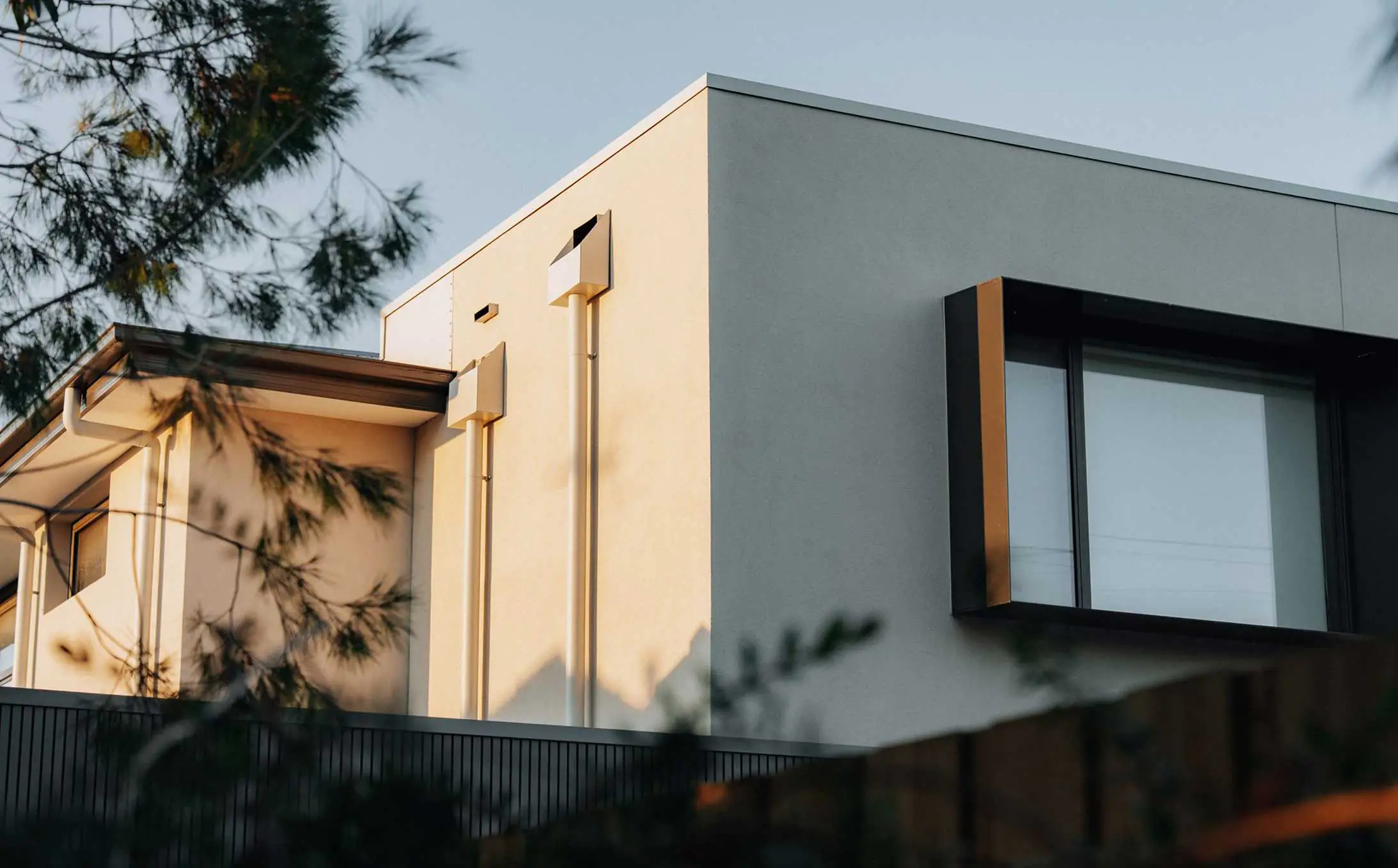 Modern minimalist house corner with beige and gray walls, a large square window, and tree branches in the foreground.