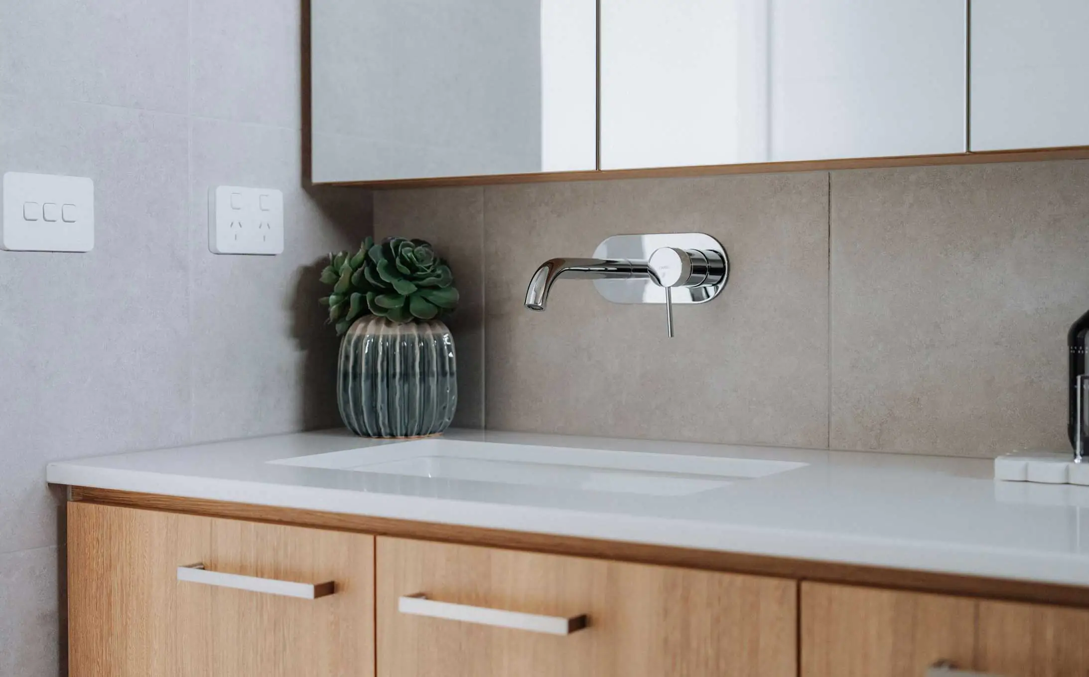 Modern kitchen sink area with a chrome wall-mounted faucet, white countertop, and wooden drawers beneath; a ribbed vase with a succulent sits on the counter.