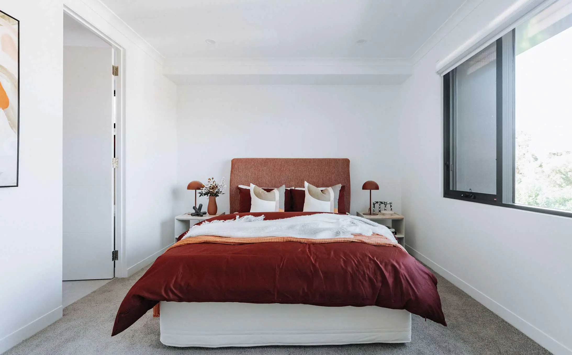 Bedroom with an orange-headboard bed, burgundy duvet, white walls, and a large window to the right.
