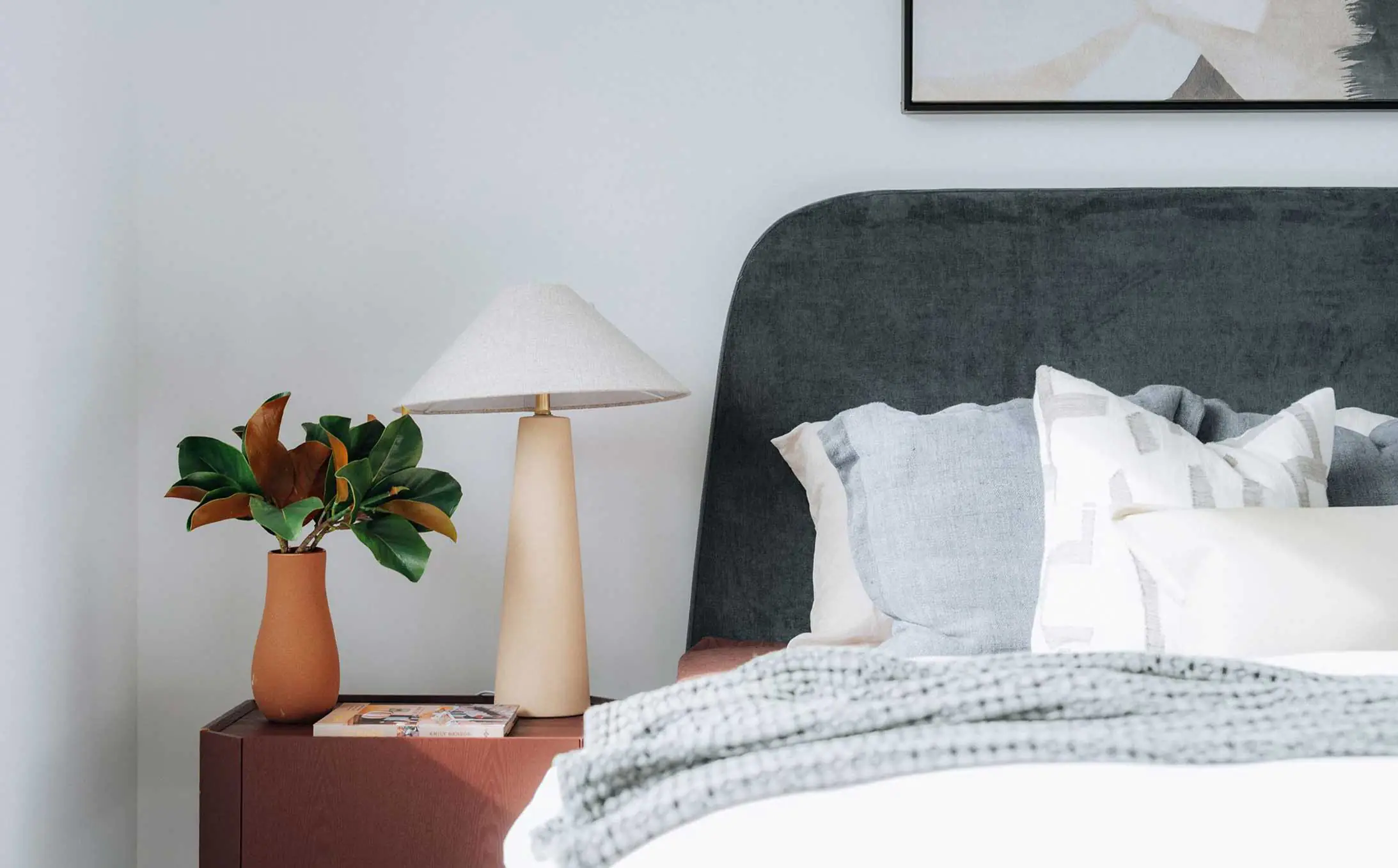 Corner bedroom scene with a wooden nightstand featuring a plant, a beige lamp, and a magazine beside a gray upholstered headboard and pillows.