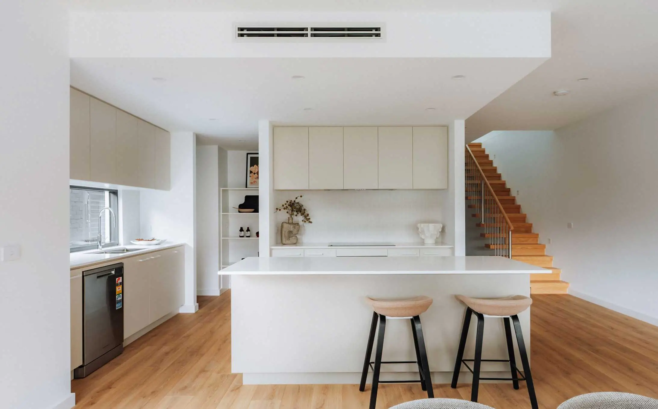 Open-concept kitchen with white cabinets, a large white island, and three black-framed bar stools; wooden stairs visible to the right.
