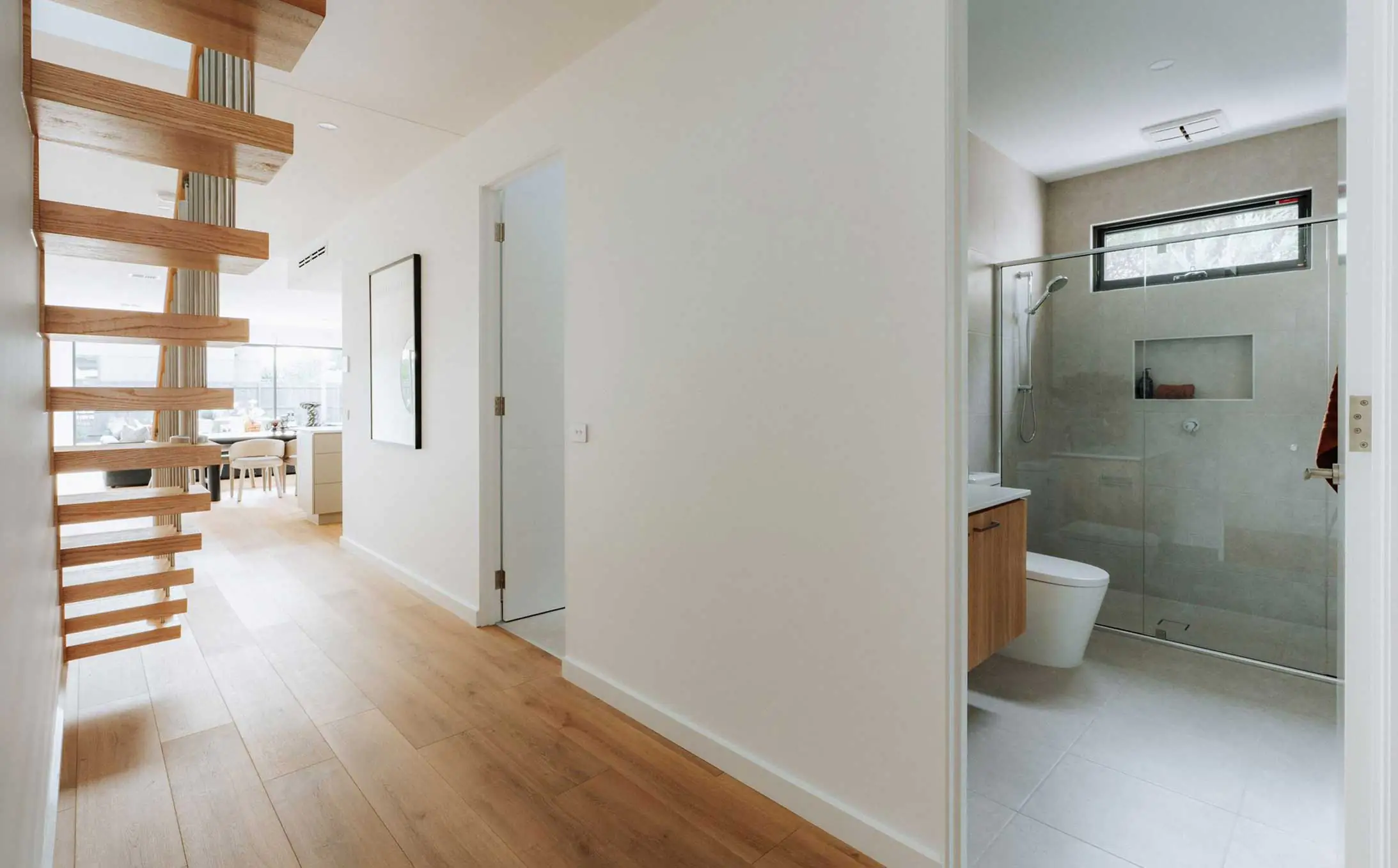 Bright modern hallway with a floating wooden staircase on the left and a glass-enclosed bathroom to the right, opening to a dining area in the distance.