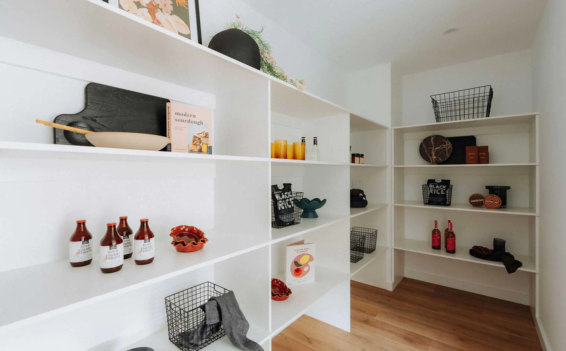 White open shelving display in a shop with bottles, bowls, and baskets arranged on multiple shelves, plus a cookbook and decorative items on top shelf
