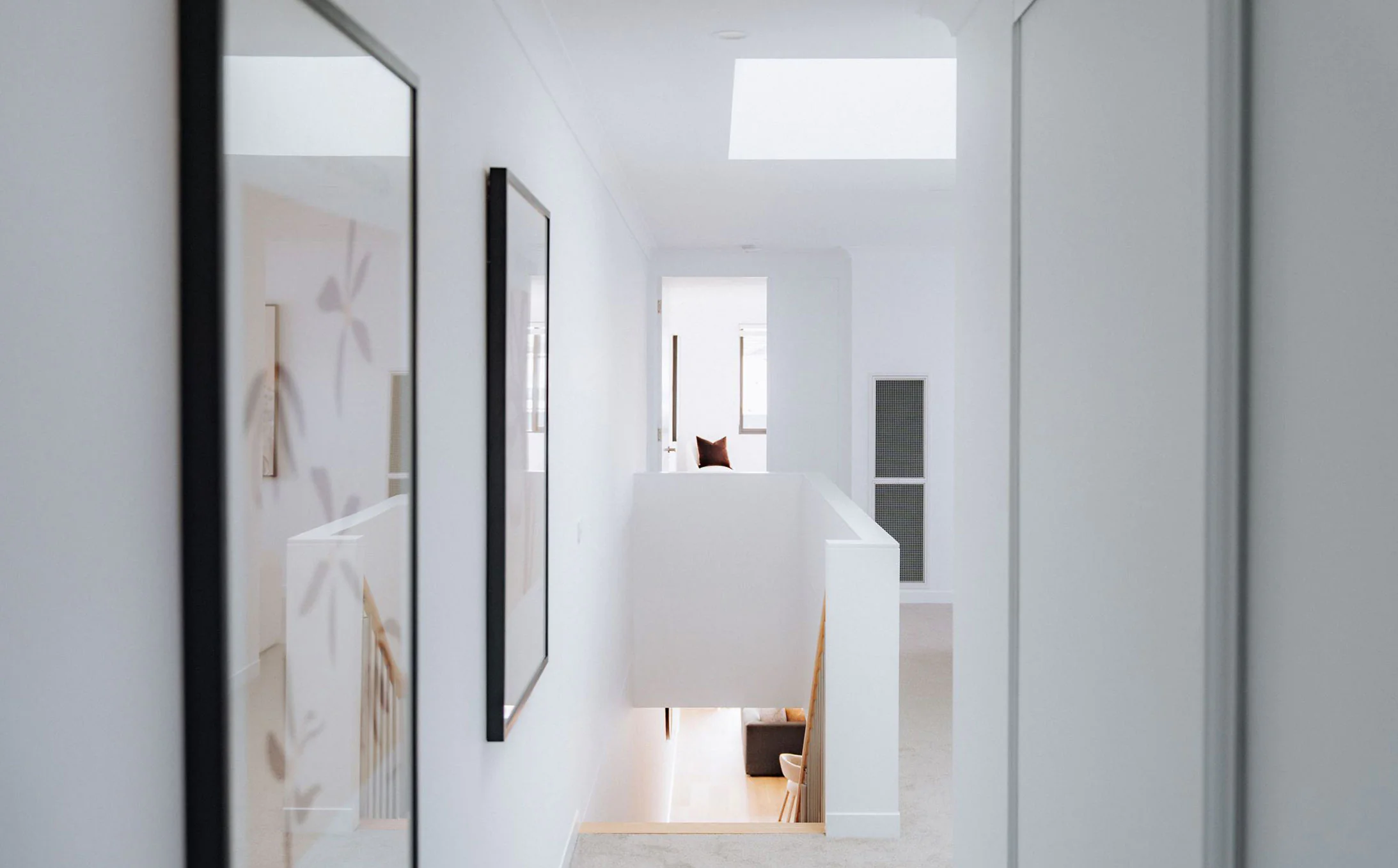 Bright white hallway with mirrored wall on the left, leading to a sunlit living area with a brown cushion in view.