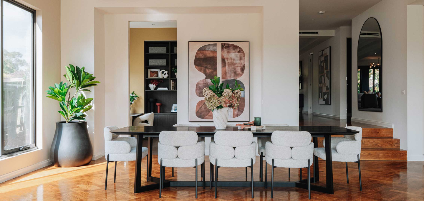 Bright dining room with a long dark table, eight white upholstered chairs, a vase of flowers, and a large window with a tall plant nearby.